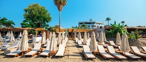 A beach with rows of sun loungers and umbrellas, a palm tree, and a building in the background.