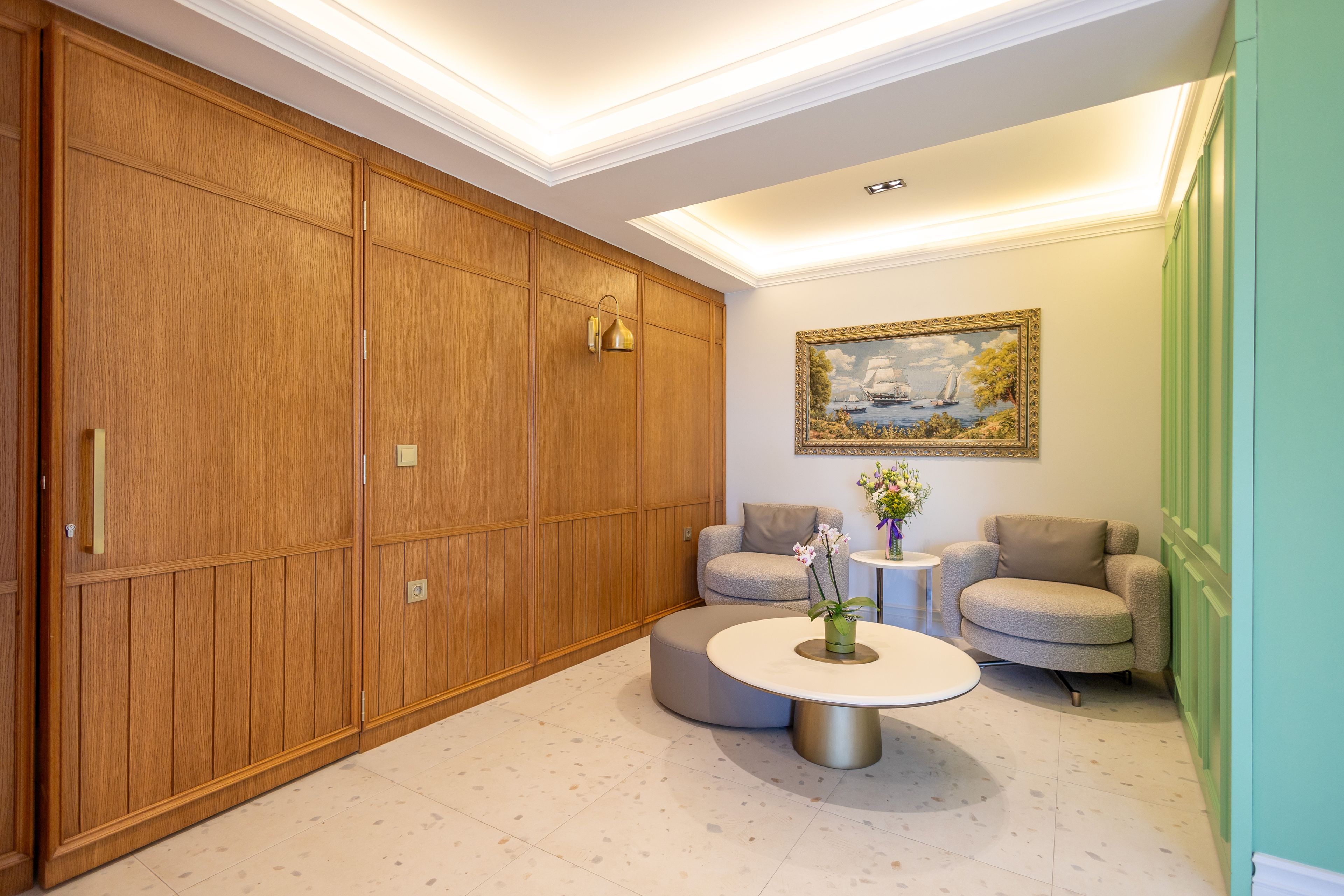 A room with a large wooden wardrobe, two armchairs, a round coffee table, and a framed painting on the wall.