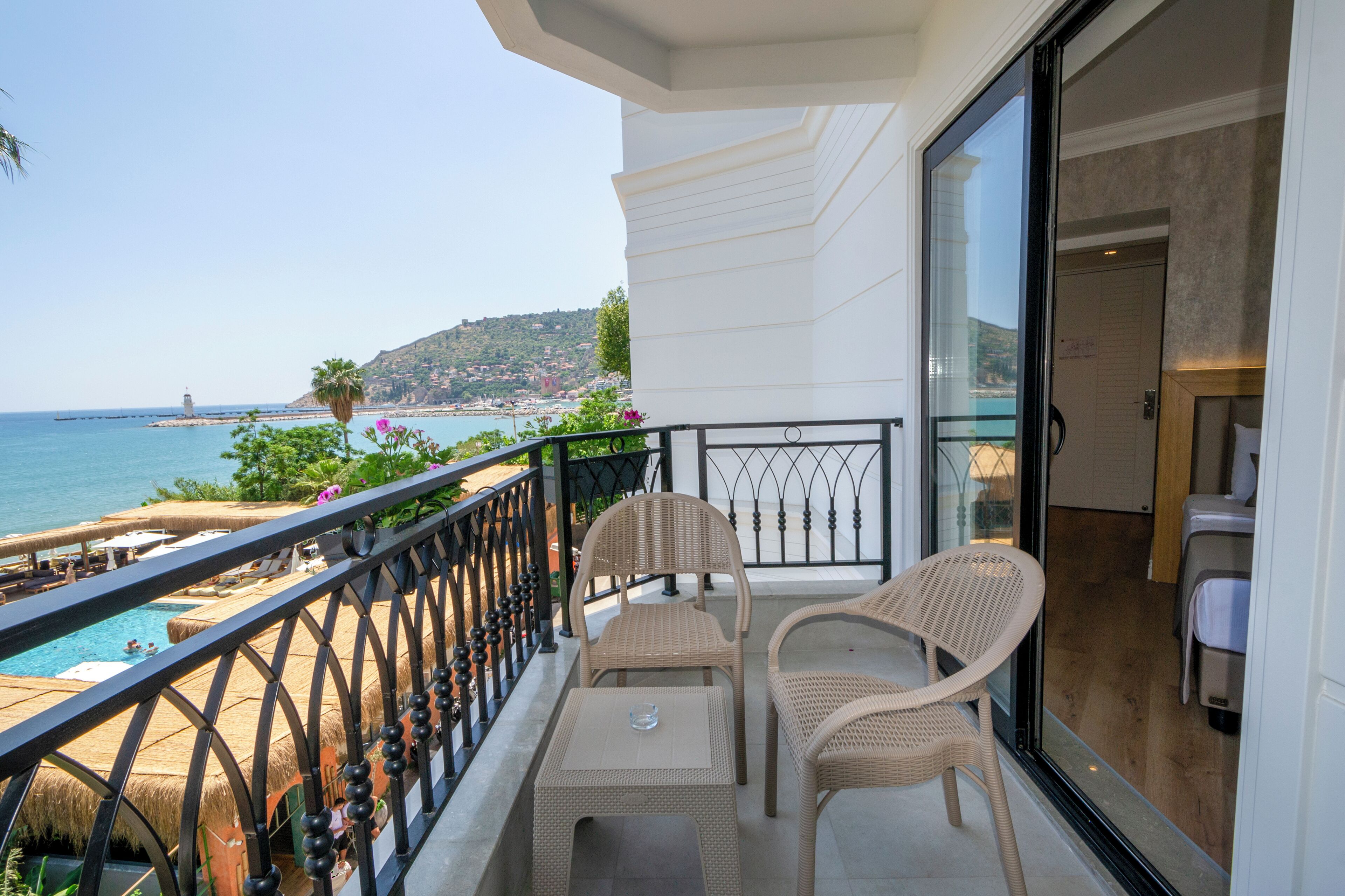 A balcony with wicker furniture, a glass door leading to a room, and a view of the sea and hills.