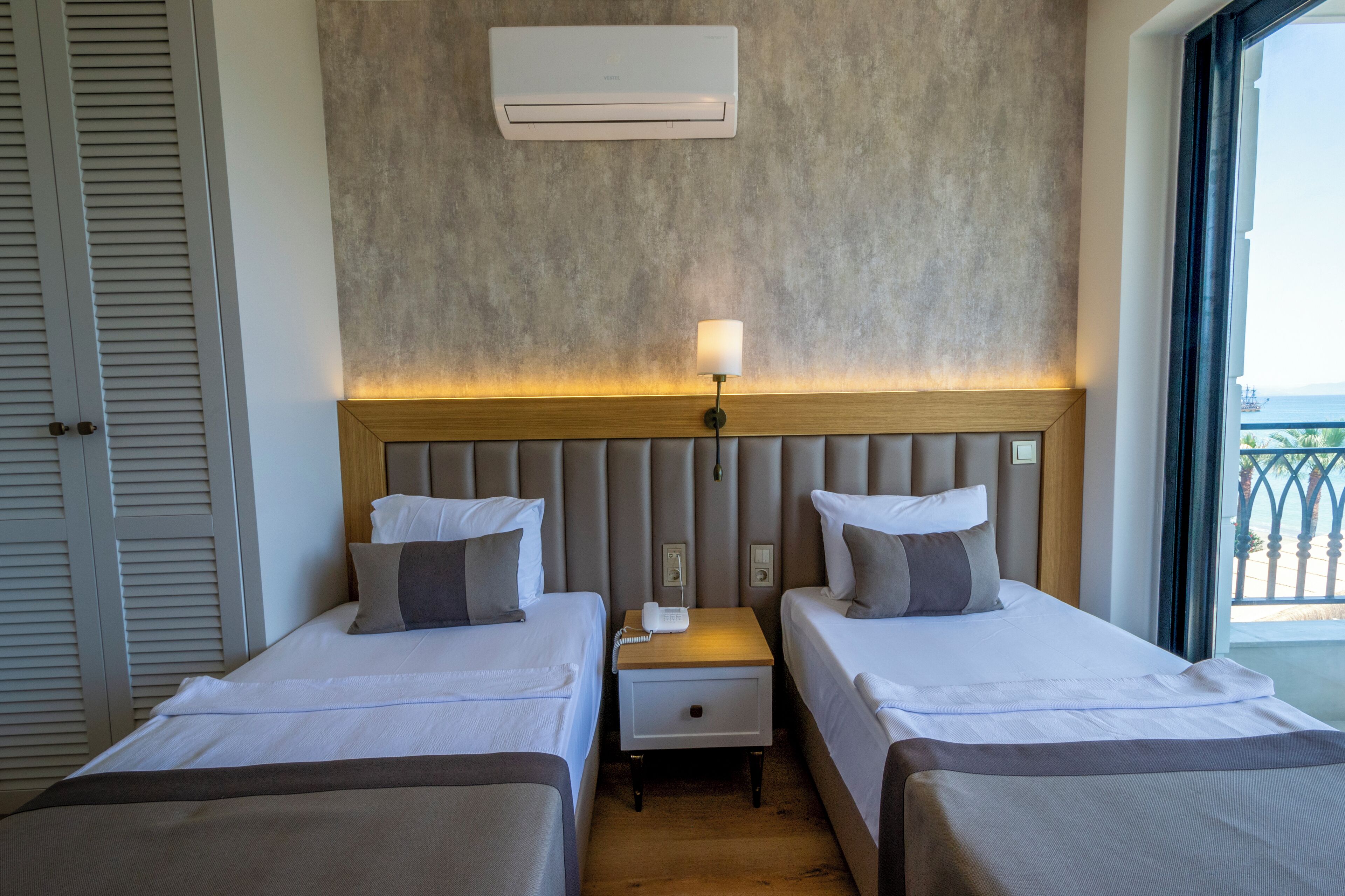 A hotel room with two beds, a nightstand with a lamp, an air conditioner, and a view of the outdoors.