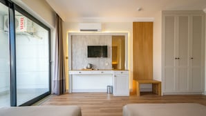 Ein modernes Hotelzimmer mit einer Schiebeglasdoor, einer Küchenzeile inklusive Fernseher, einer hölzernen Bank und einem Kleiderschrank.