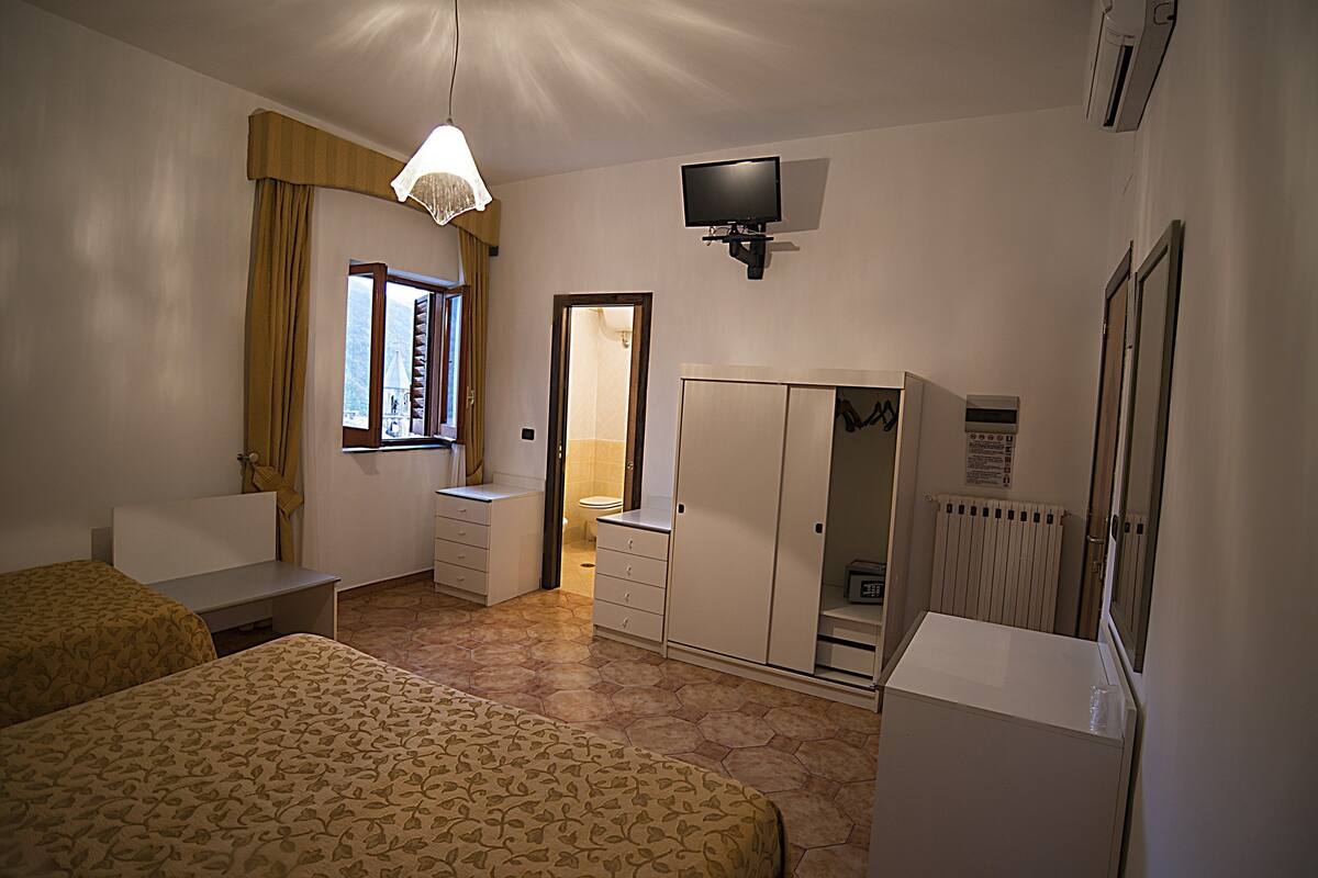A hotel room with two beds, a desk, a wardrobe, a television, and a window with curtains.