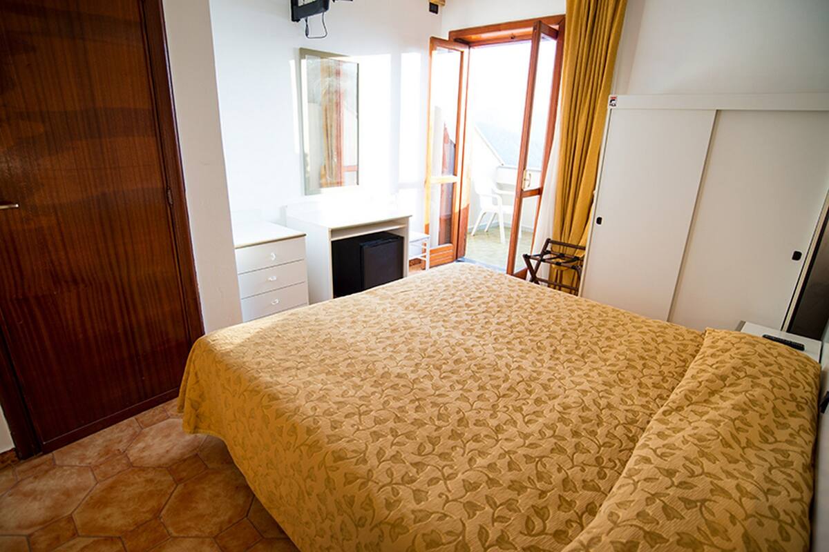 A bedroom with a large bed, a desk, and a chair. There is a sliding glass door leading to a balcony.