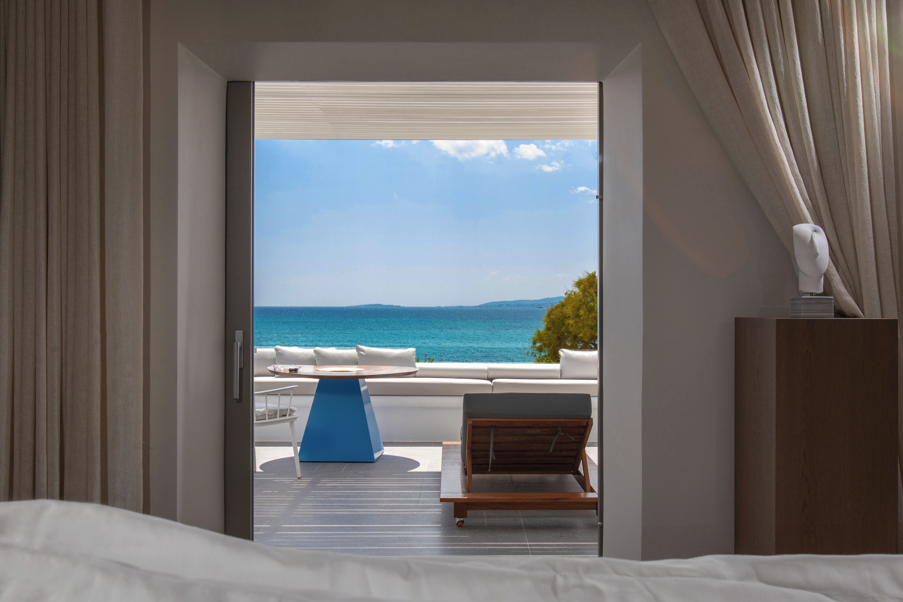 A room with a bed, a view of the sea, a blue chair, and a wooden cabinet.