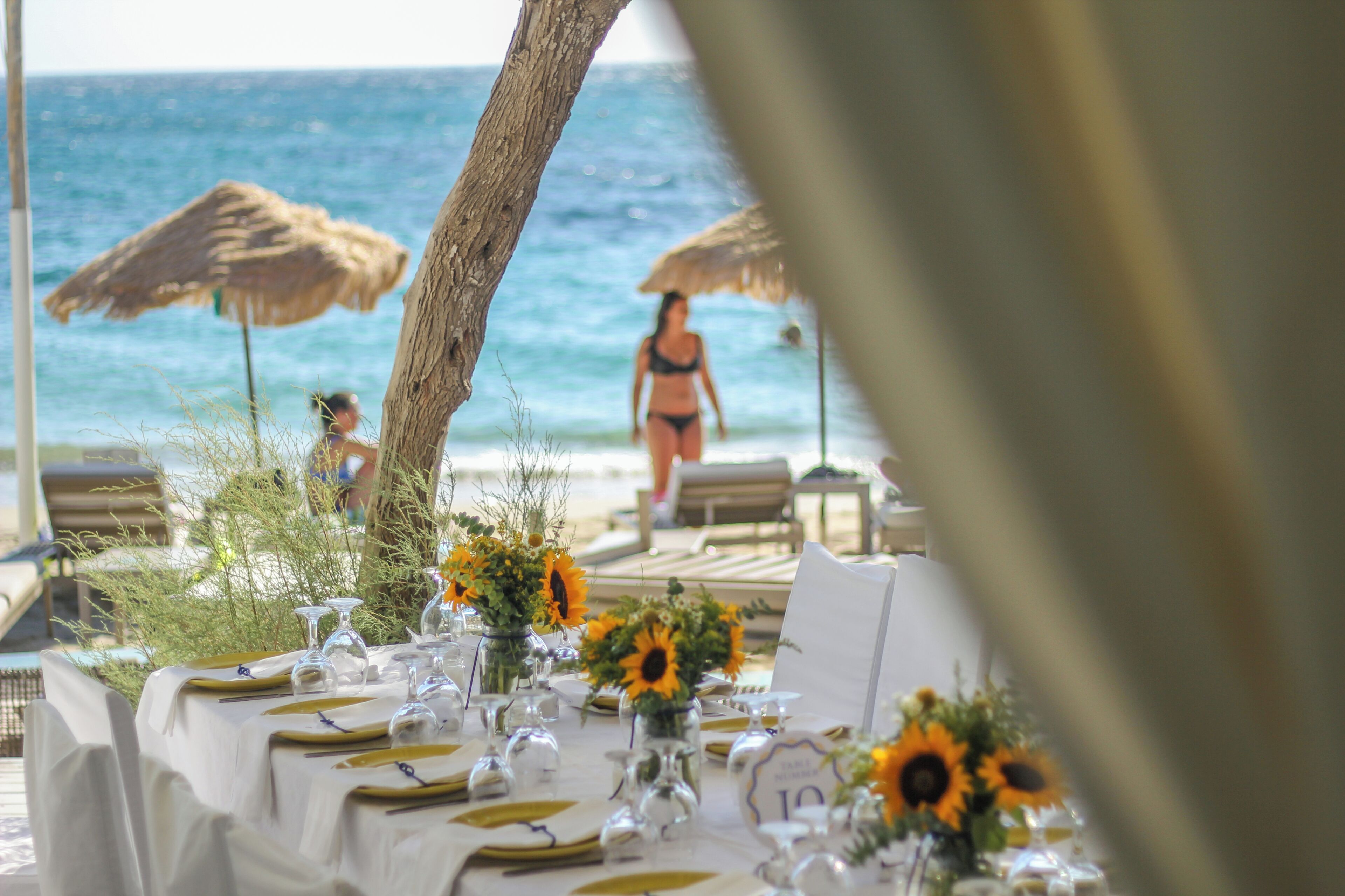 A beachside dining table set with sunflowers, clear glassware, and numbered place settings.
