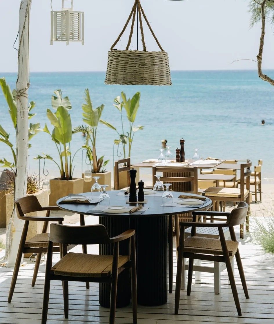 An outdoor dining area with a view of the sea, featuring round tables, chairs, and hanging lanterns.