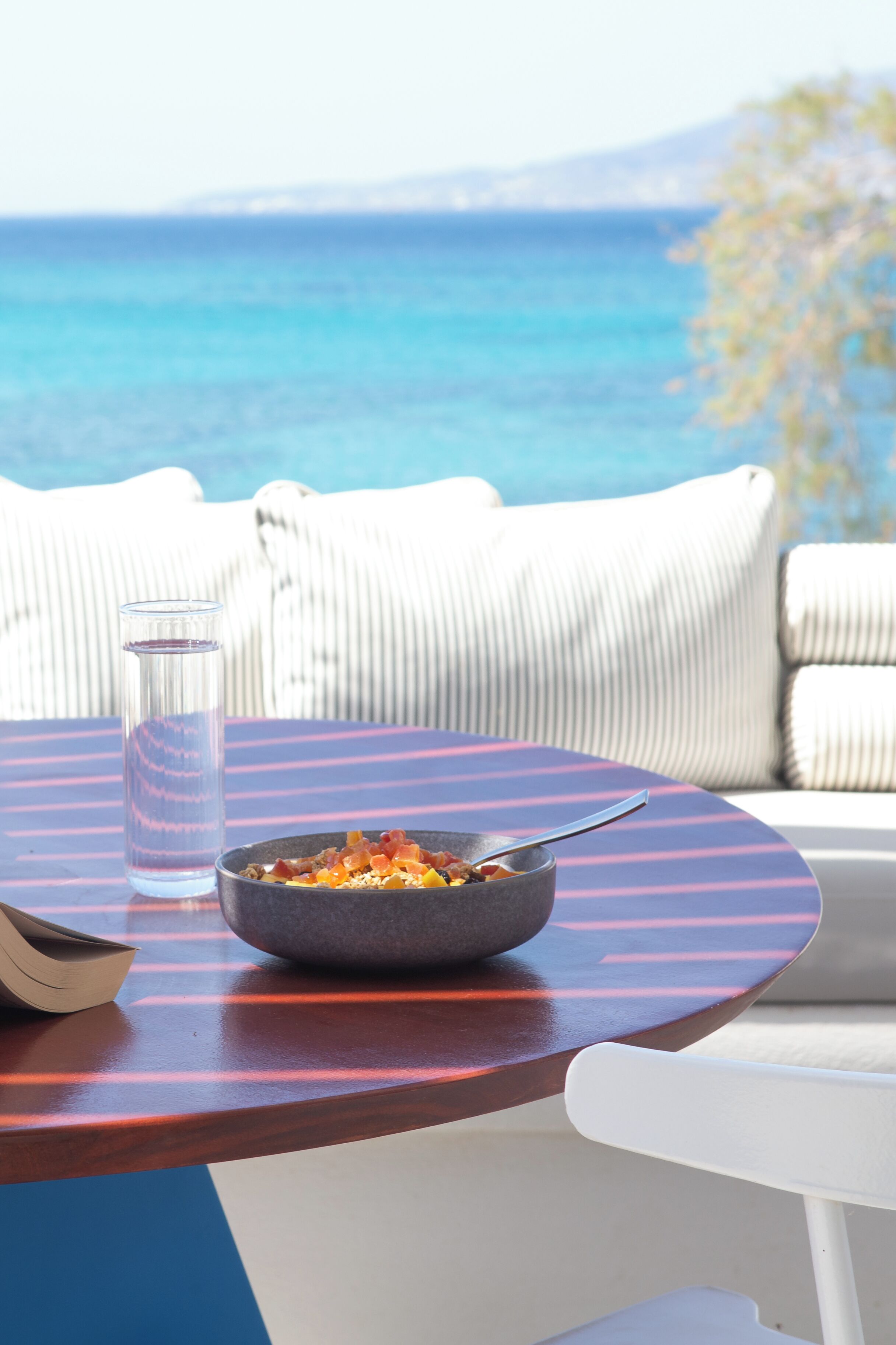 A round table with a bowl of food, a glass of water, and a spoon. The table is on a balcony with a view of the sea.
