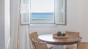 Une table ronde en bois, quatre chaises en osier, une lampe blanche et une vue sur la mer par une fenêtre ouverte.