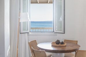 Uma mesa de madeira redonda com quatro cadeiras de vime, um abajur branco e vista para o mar através de uma janela aberta.