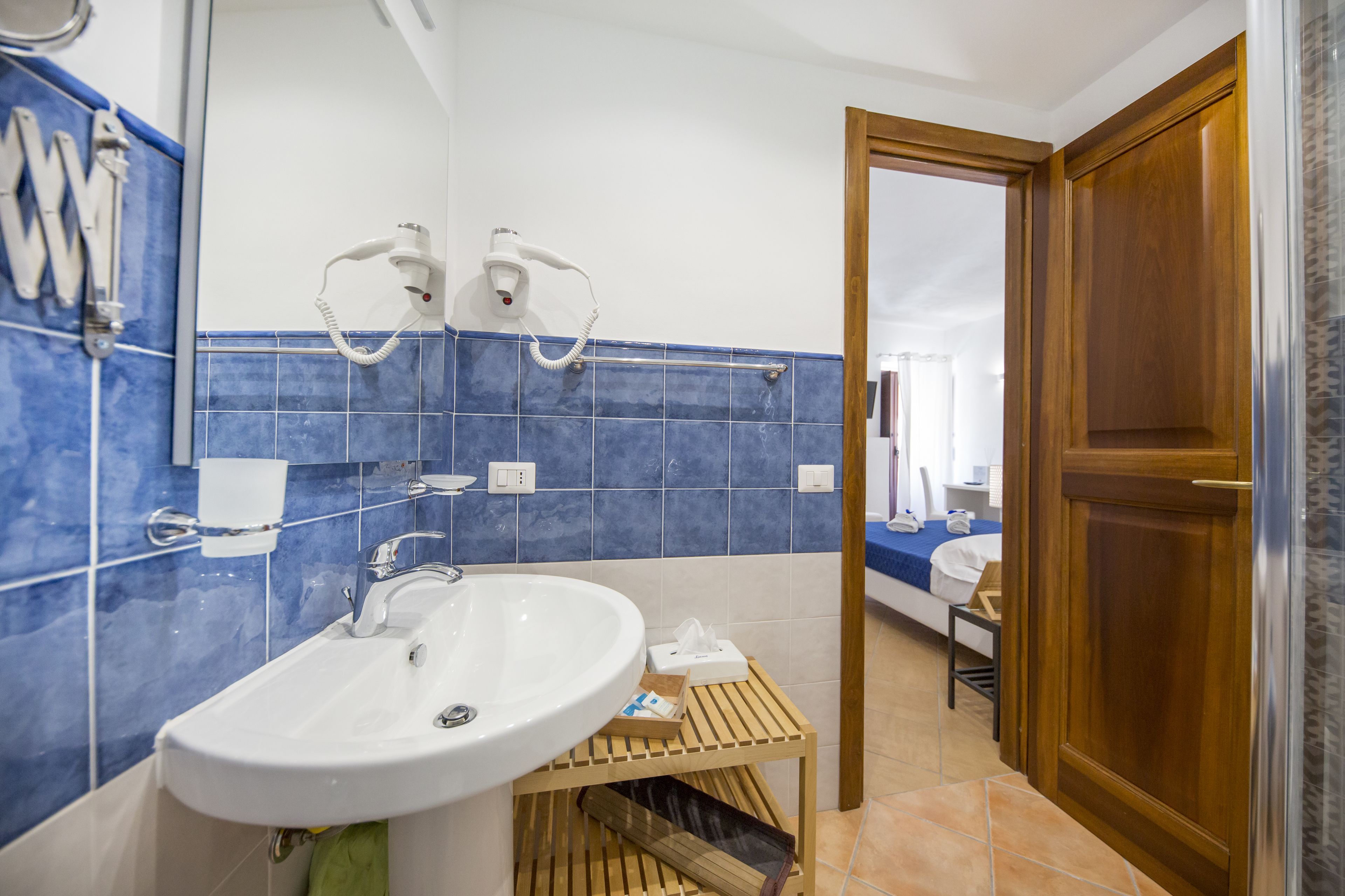 A bathroom with a sink, two hair dryers mounted on the wall, a wooden shelf with toiletries, and a door leading to another room.