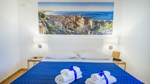 Habitación de hotel con una cama grande, dos almohadas blancas, una colcha azul y dos toallas enrolladas con cintas azules.
