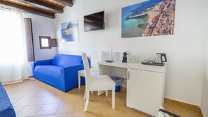 Una habitación con un sofá azul, un escritorio blanco y una silla. Hay pinturas en la pared, una ventana con cortinas y un piso de baldosas.