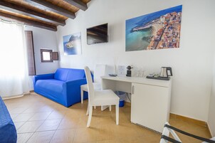 Una habitación con un sofá azul, un escritorio blanco y una silla. Hay pinturas en la pared, una ventana con cortinas y un piso de baldosas.