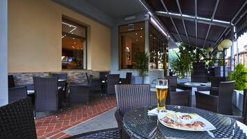 Un patio al aire libre con mobiliario de mimbre, una mesa redonda con un plato de comida y una cerveza, y vista del interior a través de una puerta de cristal.