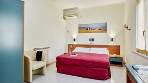 A hotel room with a bed, a chair, a desk, and a painting on the wall.