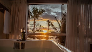 Una habitación con bañera, ventana con cortinas y vistas a palmeras y un atardecer.