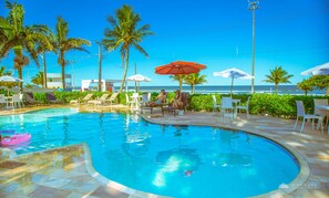 Une piscine entourée de chaises longues et de parasols, bordée de palmiers et offrant une vue sur l’océan.