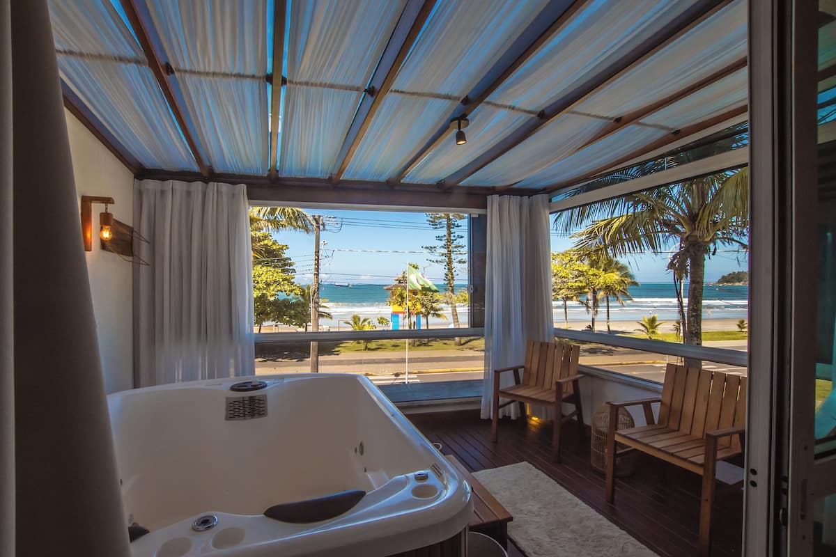 Une pièce dotée d’une grande fenêtre donnant sur une plage, d’une terrasse en bois et d’un jacuzzi.