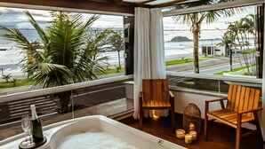 Un cuarto con una bañera grande llena de burbujas, sillas de madera, un ventanal con vista a palmeras y a la playa, y una botella de champán al lado de la bañera.