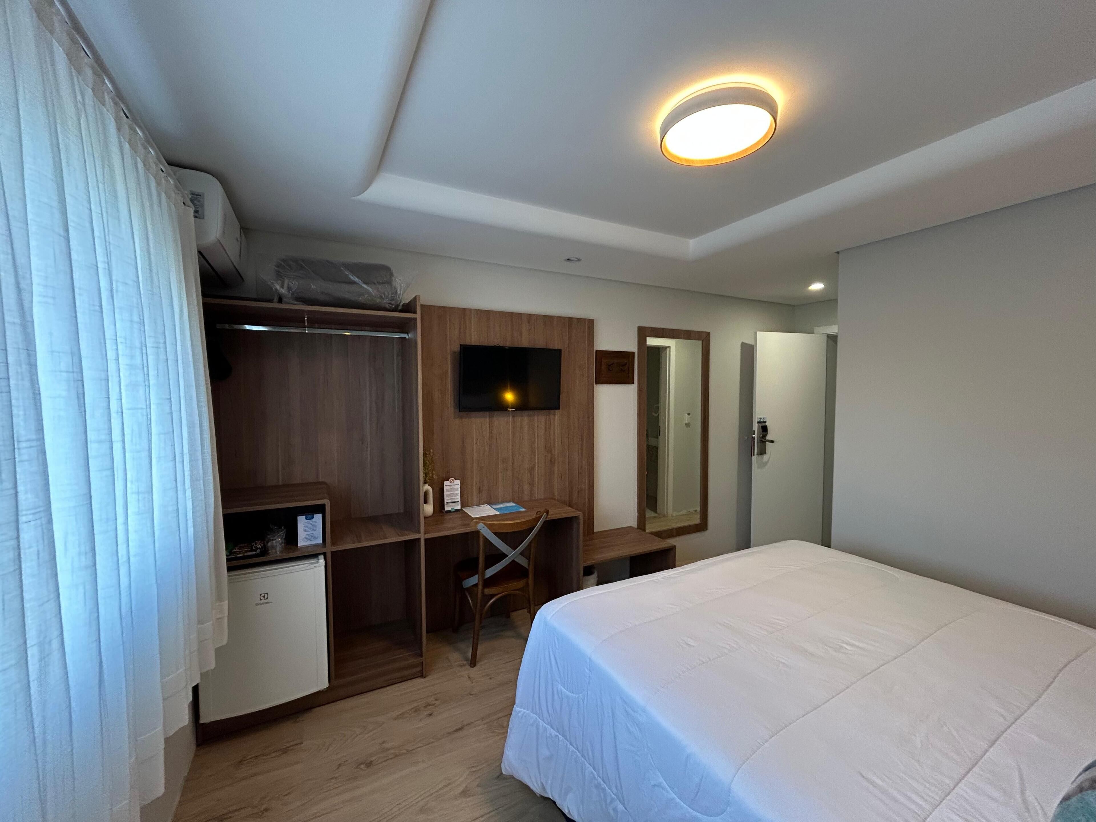 Habitación de hotel con cama, escritorio, silla, armario, televisor y espejo.