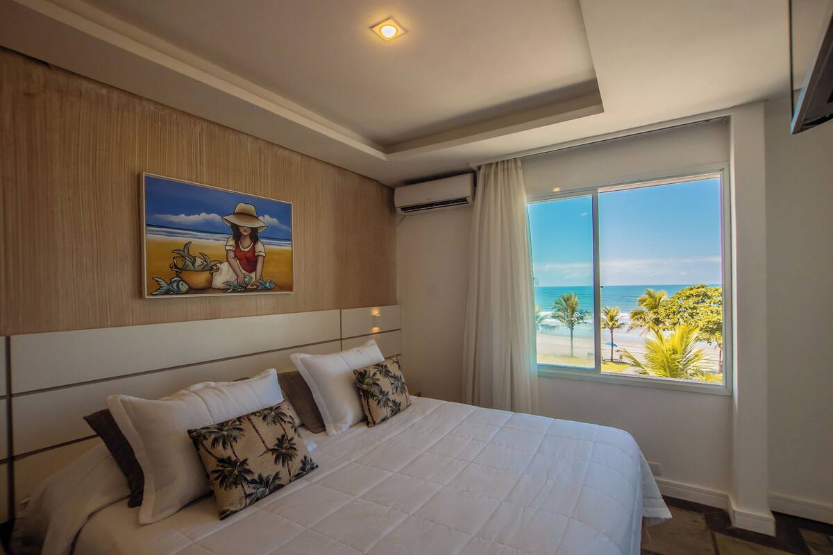 Une chambre d’hôtel avec un grand lit, un tableau représentant une scène de plage, un appareil de climatisation et une fenêtre donnant sur la plage.