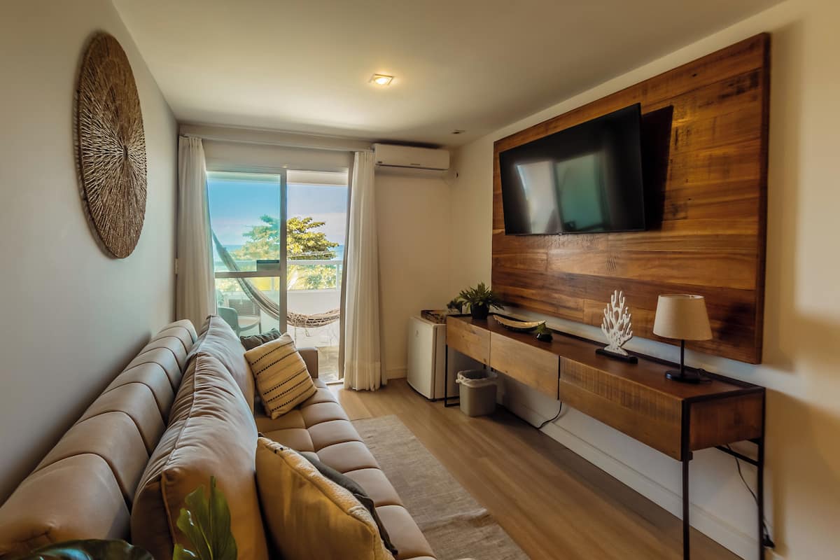 Un salon moderne avec un canapé, une console en bois, un téléviseur fixé au mur et une vue sur l’extérieur.