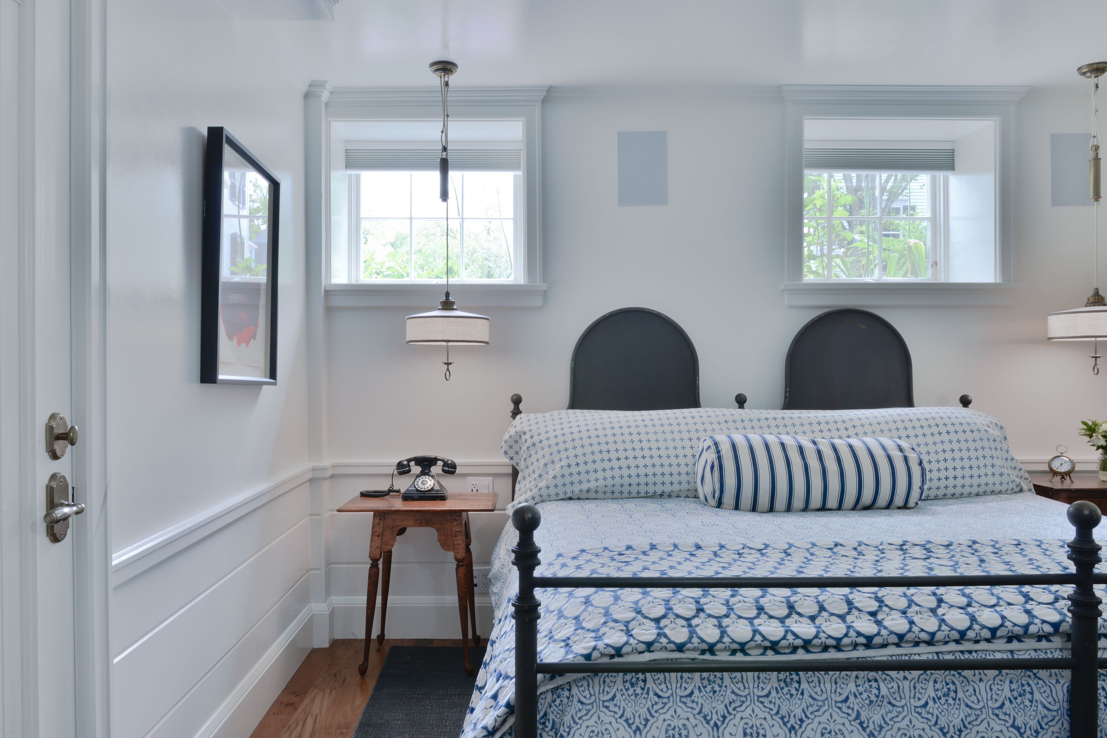A bedroom with a bed, two windows, a bedside table with a lamp, and a framed picture on the wall.