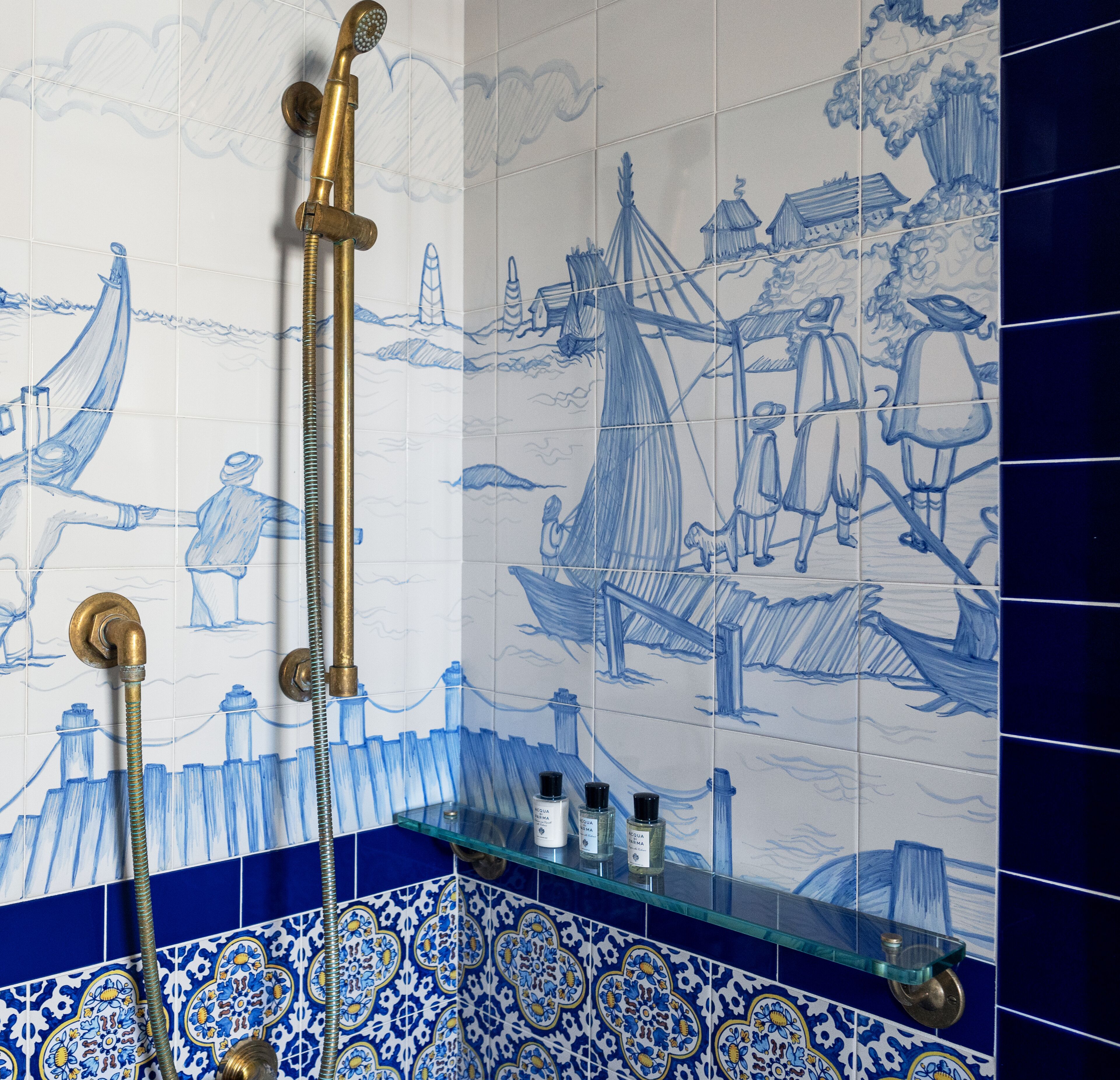 A bathroom with a blue and white tiled wall featuring maritime scenes, a glass shelf with toiletries, and a brass showerhead.