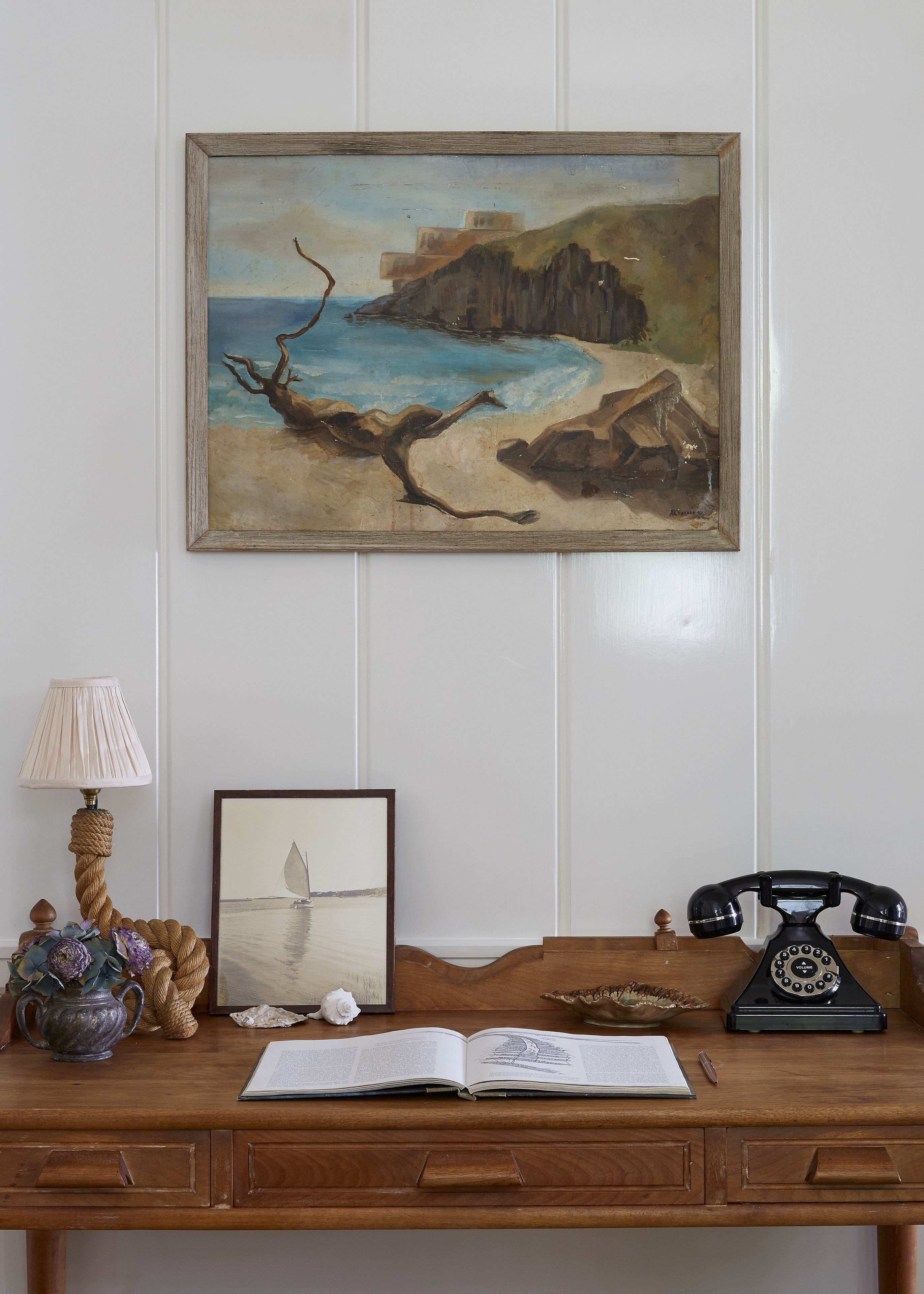 A desk with a book, a lamp, a telephone, and a framed painting of a coastal scene.
