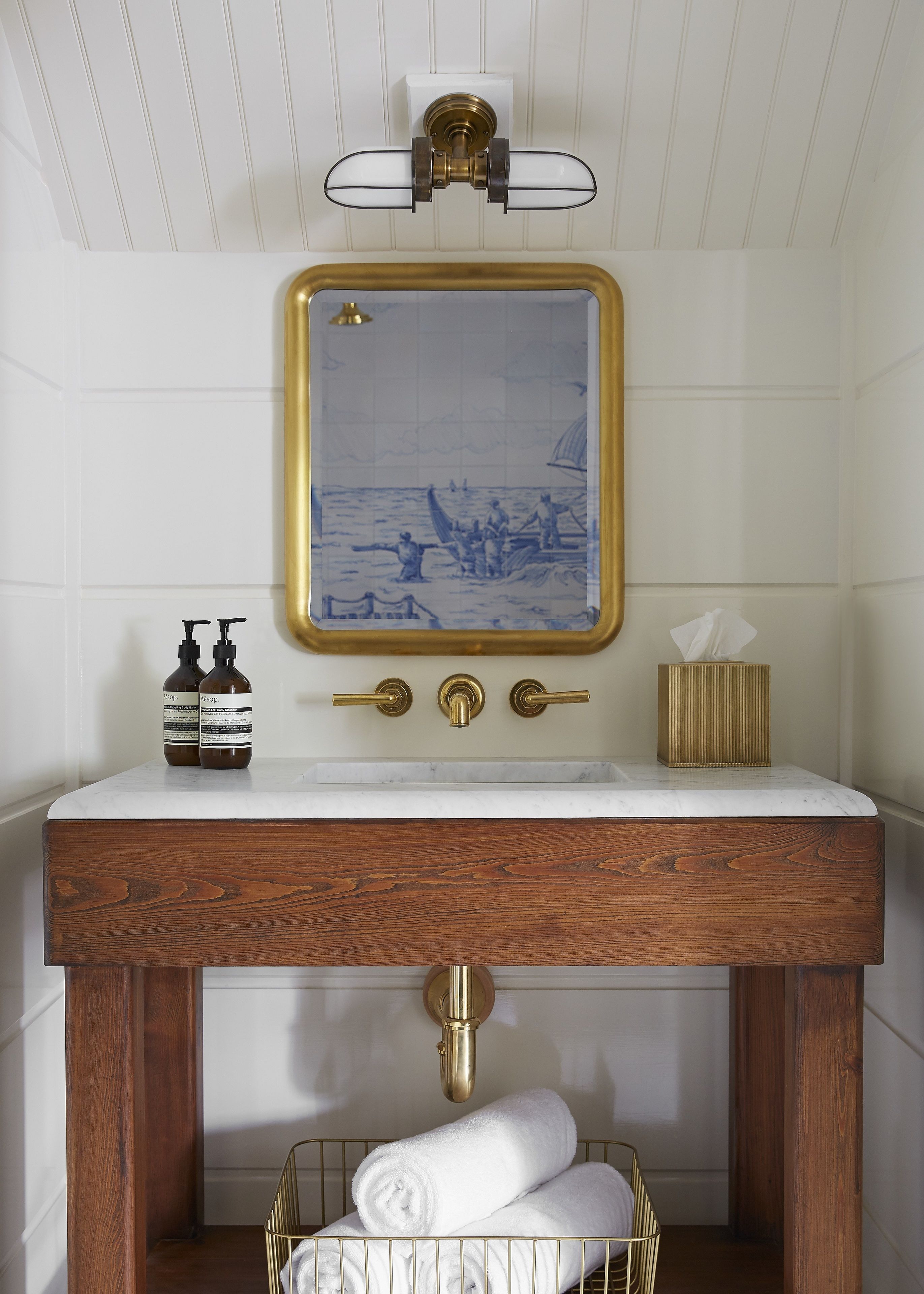 Un baño con pared de azulejos blancos, un mueble de tocador de madera con encimera de mármol, un espejo con marco dorado y un perchero con toallas enrolladas.