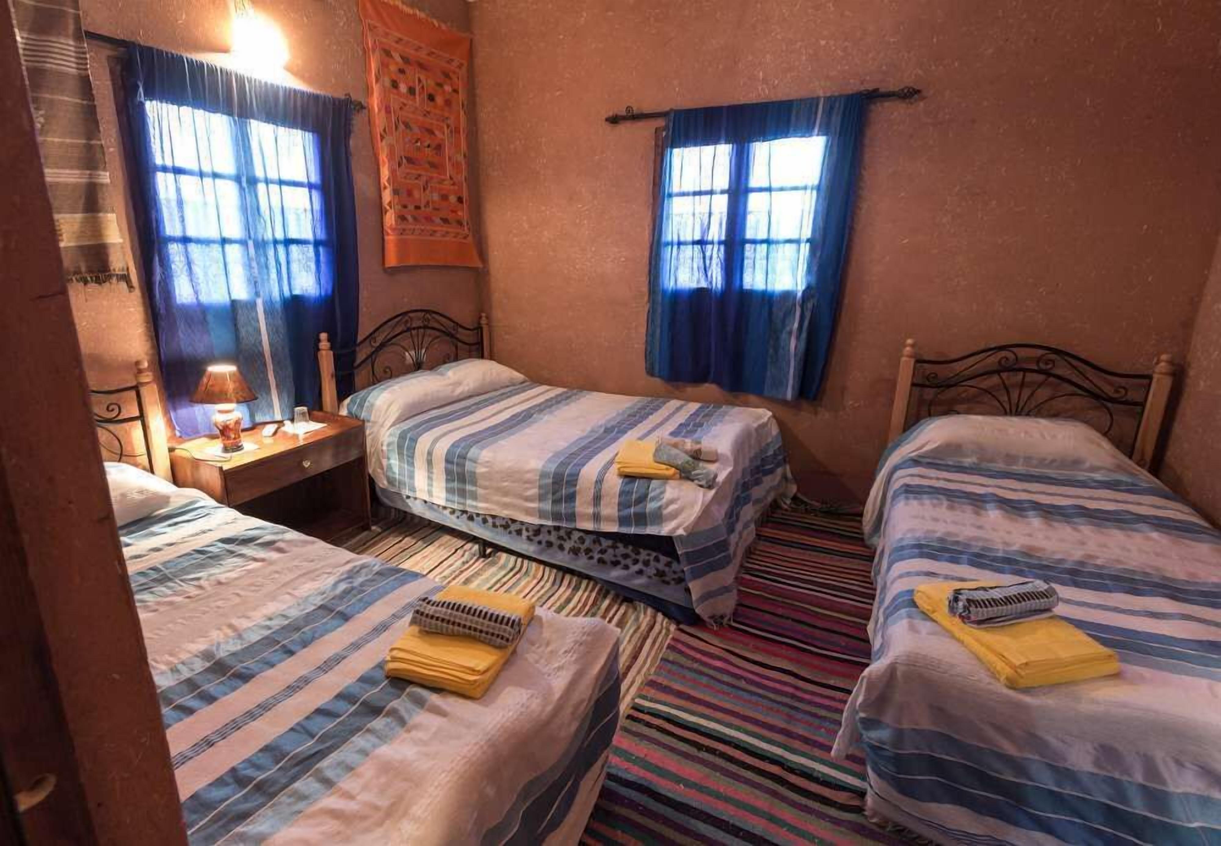 A room with three beds, each with striped bedding, a small table with a lamp, and two windows with blue curtains.