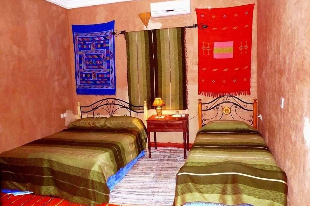 A room with two single beds, a small table with a lamp, and a window with curtains.