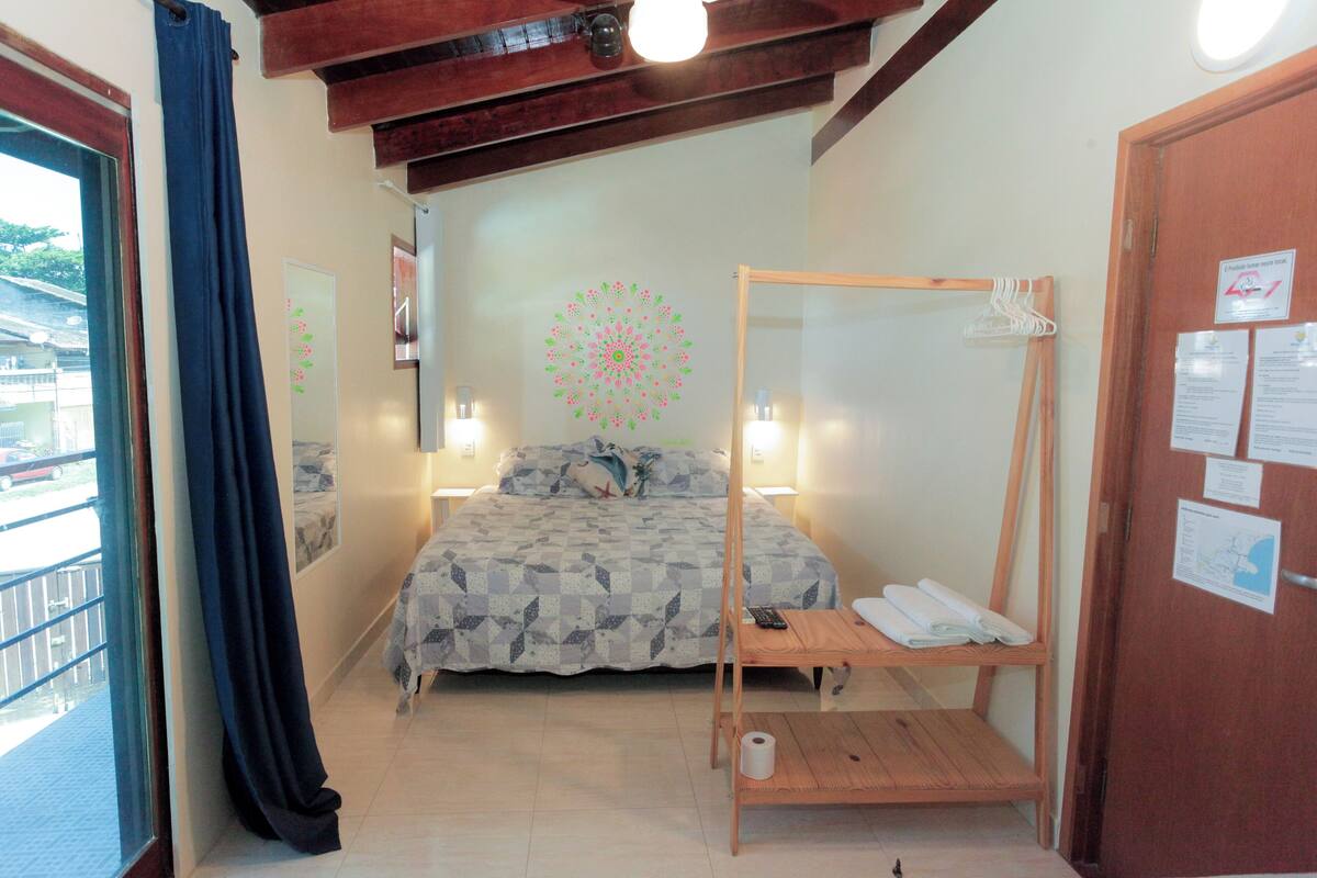 A bedroom with a bed, a wooden bedside table, a hanging chair, a window with curtains, and a door with a notice.