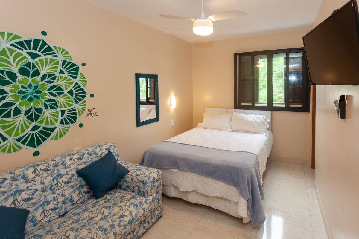 A bedroom with a bed, a sofa, a ceiling fan, a window, and a wall decoration.