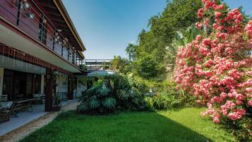 Um hotel com área de estar coberta ao ar livre, um jardim com diversas plantas e uma varanda com plantas pendentes.
