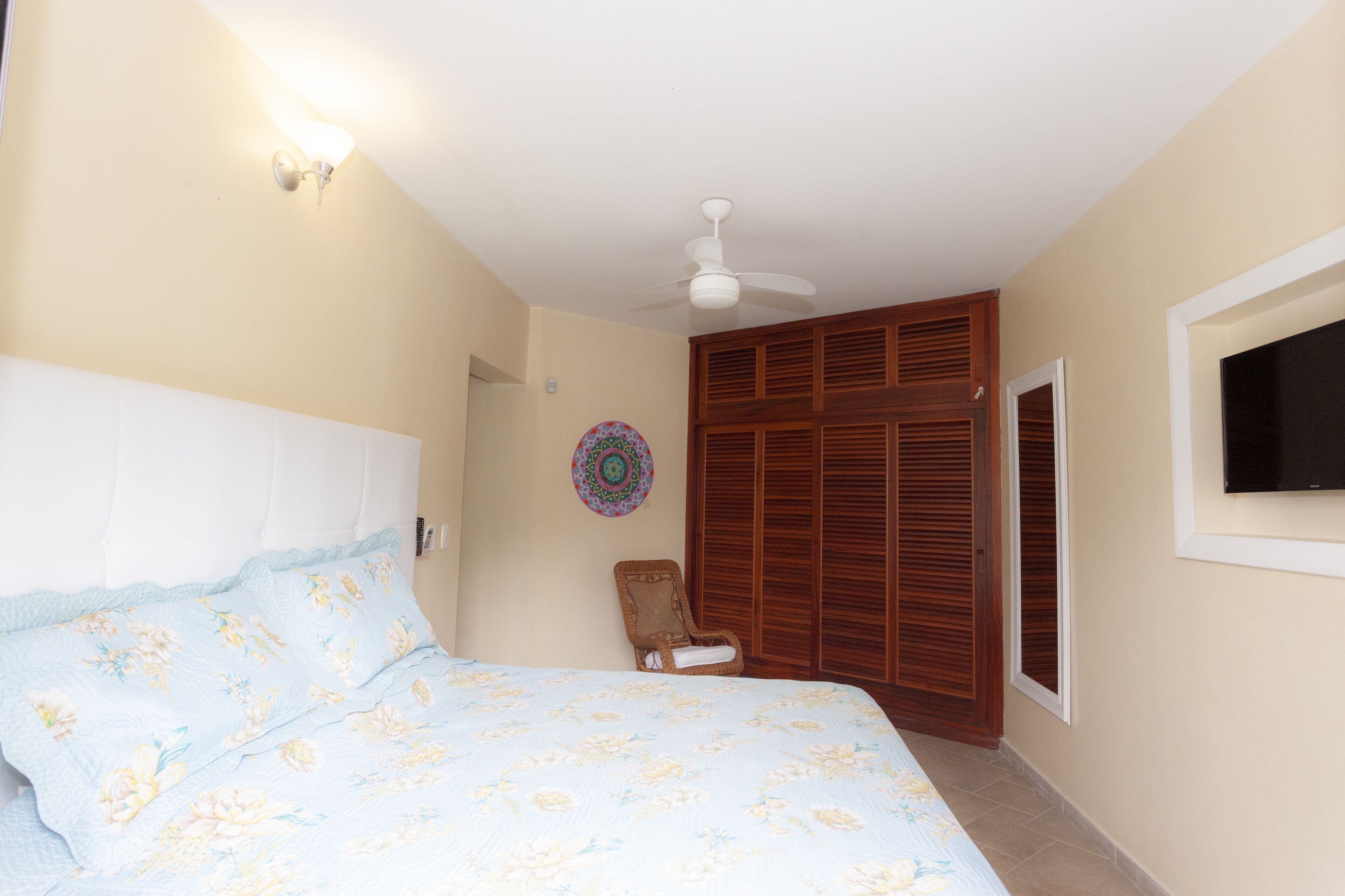 Quarto com cama, ventilador de teto, guarda-roupa de madeira e televisão fixada na parede.