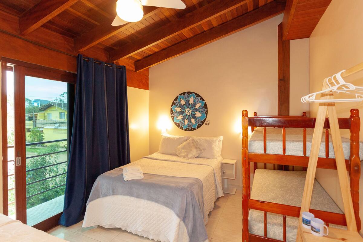 A bedroom with a bunk bed, a single bed, a window with curtains, a wall clock, and a view of houses outside.