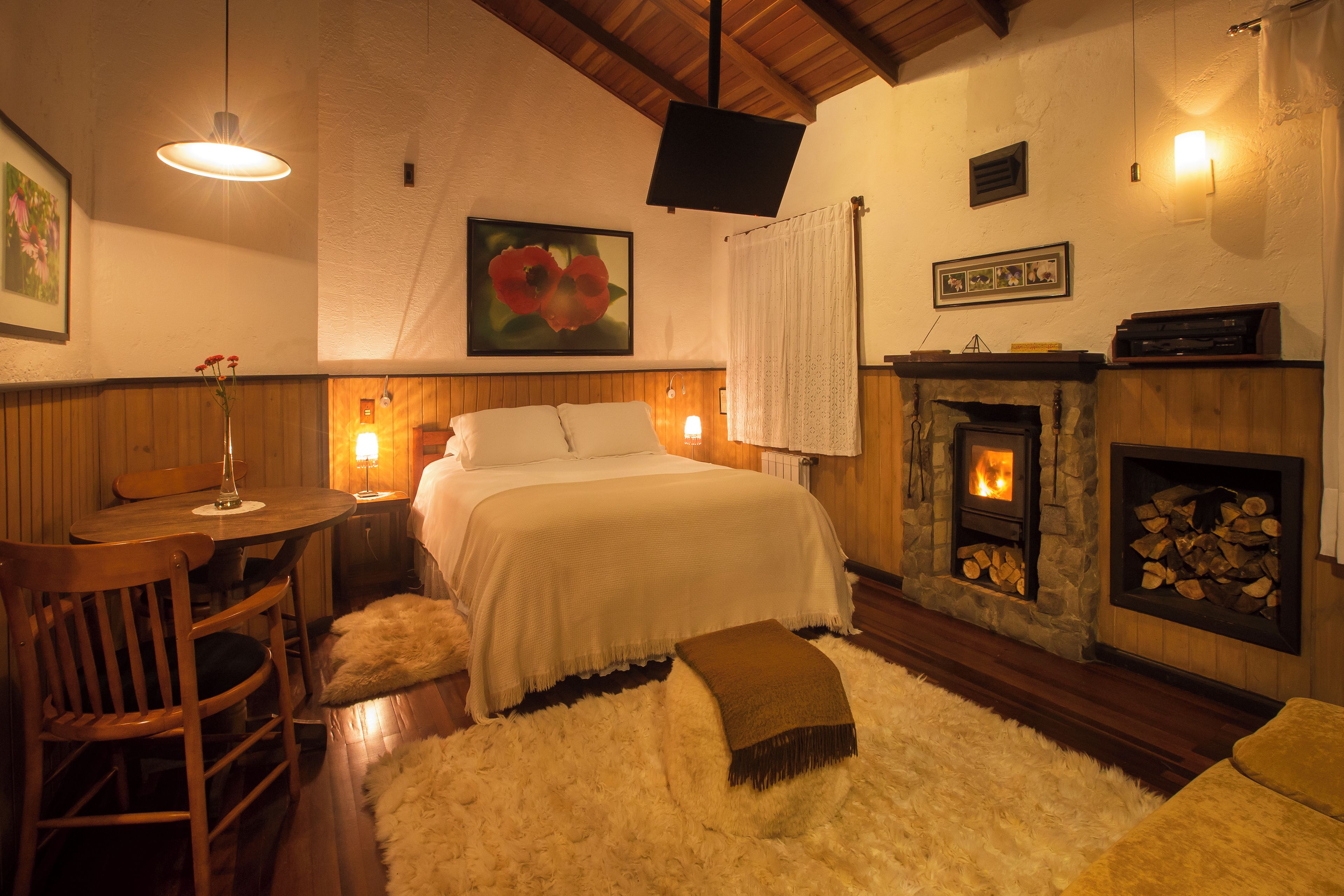 A cozy bedroom with a bed, a wooden table with chairs, a fireplace, and a painting on the wall.