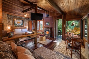 Acogedor interior de cabaña con chimenea, mobiliario de madera y un amplio ventanal con vistas a la vegetación.