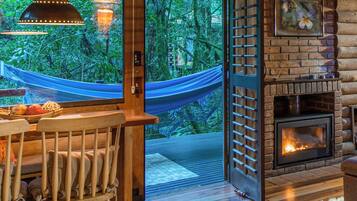 Interior acogedor de una cabaña con comedor de madera, una hamaca, chimenea y un ventanal con vistas a un bosque.