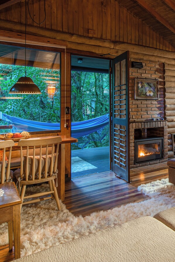 Interior acogedor de una cabaña con comedor de madera, una hamaca, chimenea y un ventanal con vistas a un bosque.