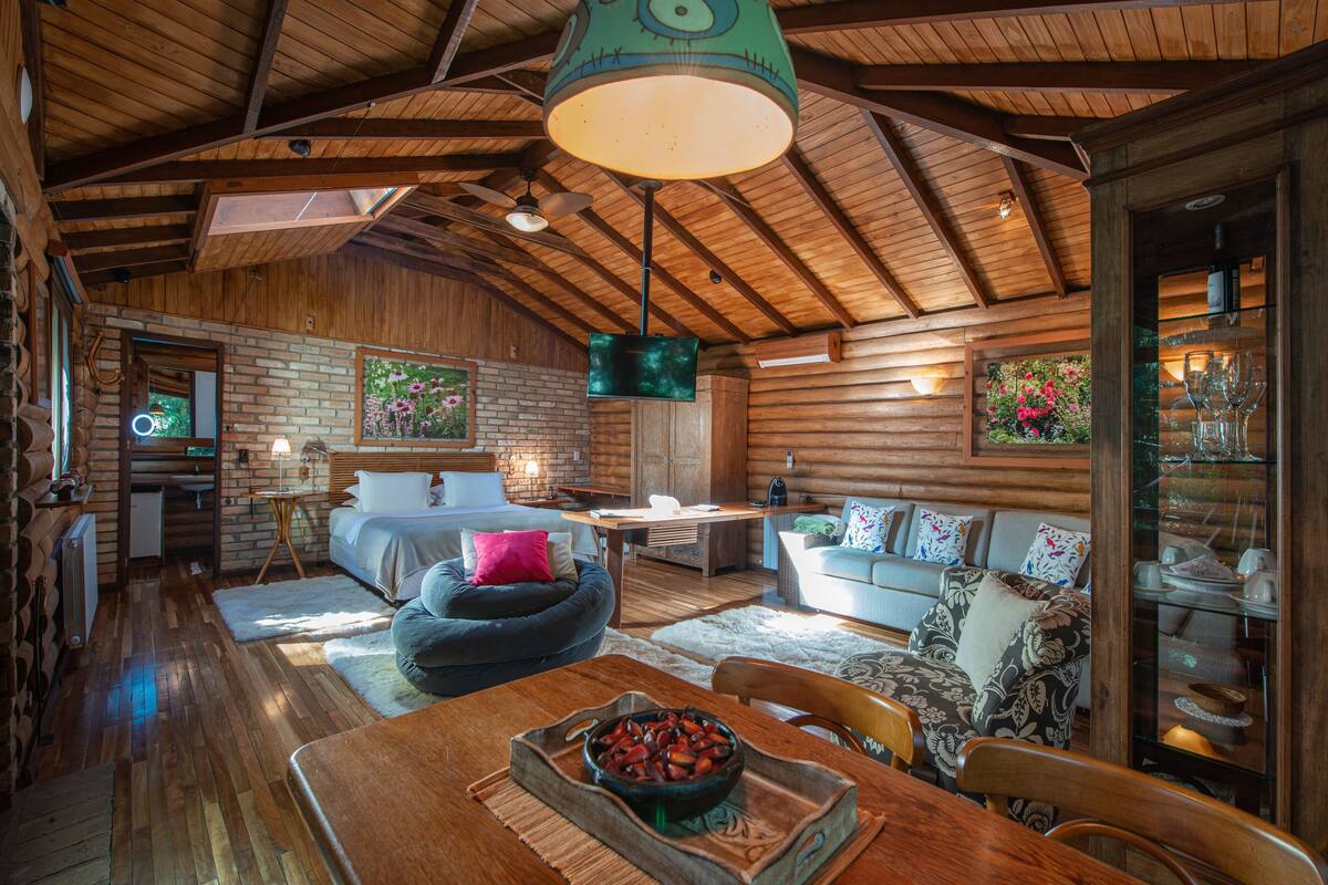 A rustic cabin interior with a wooden ceiling, a dining area with a bowl of fruit, a bed with a pink pillow, and a sofa with floral cushions.