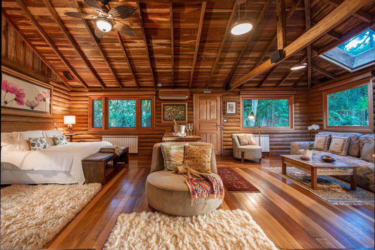 A log cabin interior with a bed, sofa, and armchair, wooden floors, and large windows.