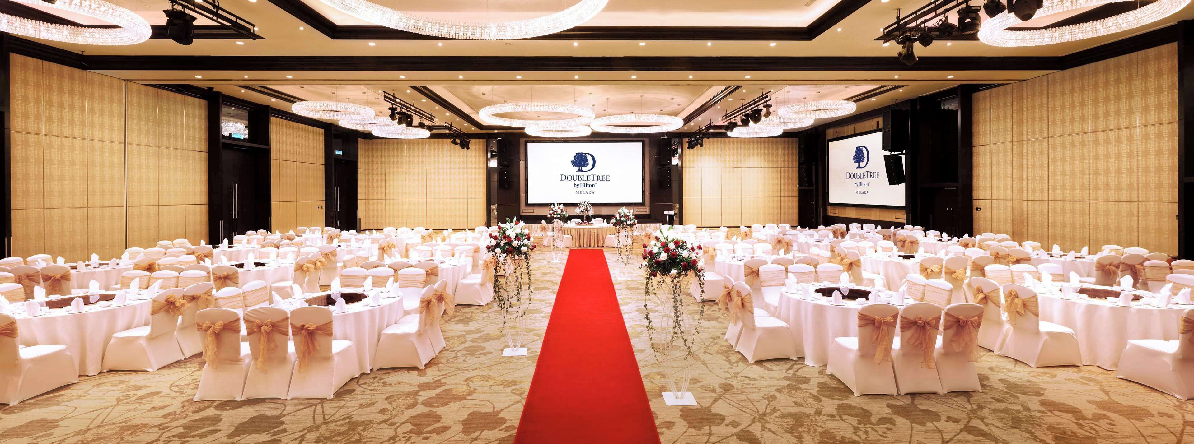 A large conference room with a red carpet, white chairs with gold sashes, and a stage with a screen displaying the DoubleTree by Hilton logo.