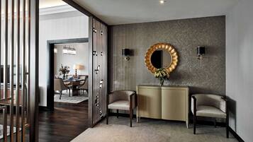 Un interior moderno con suelo de madera oscura, una consola beige, dos sillones, un espejo decorativo y una lámpara de pared.