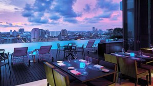 Un espace de restauration en plein air, avec des tables et des chaises, offrant une vue sur les toits d'une ville et sur une étendue d'eau au crépuscule.