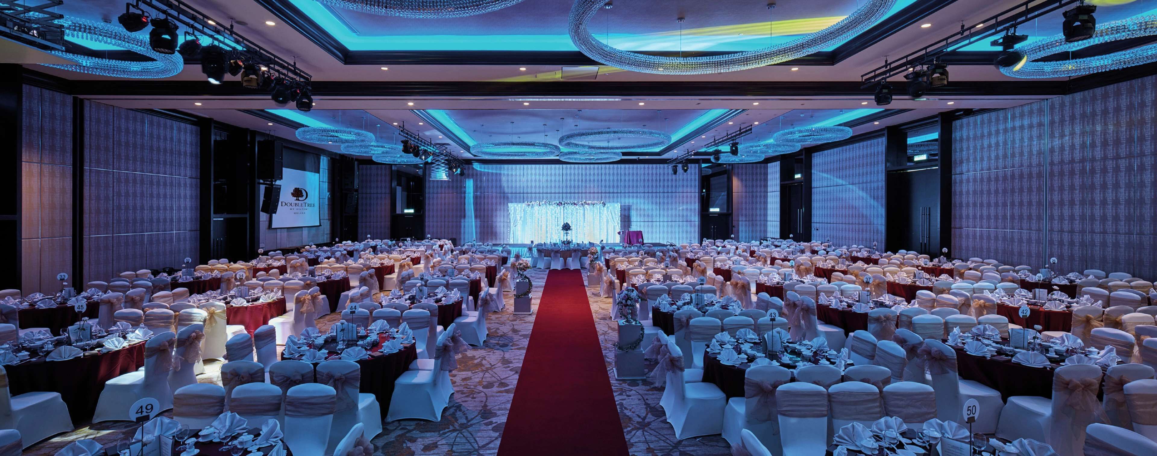 A large banquet hall with rows of tables, each with white tablecloths and chairs, and a red carpet aisle.