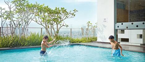 Zona de piscina en la azotea con dos personas jugando en el agua, rodeada de vegetación y un edificio moderno con grandes ventanales.
