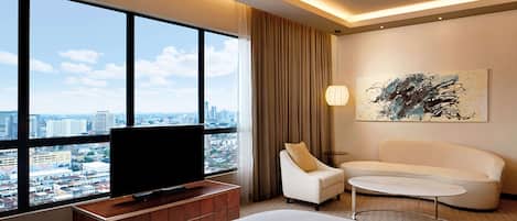 A hotel room with a large window offering a city view, a flat-screen TV, a sofa, a small table, and a painting on the wall.