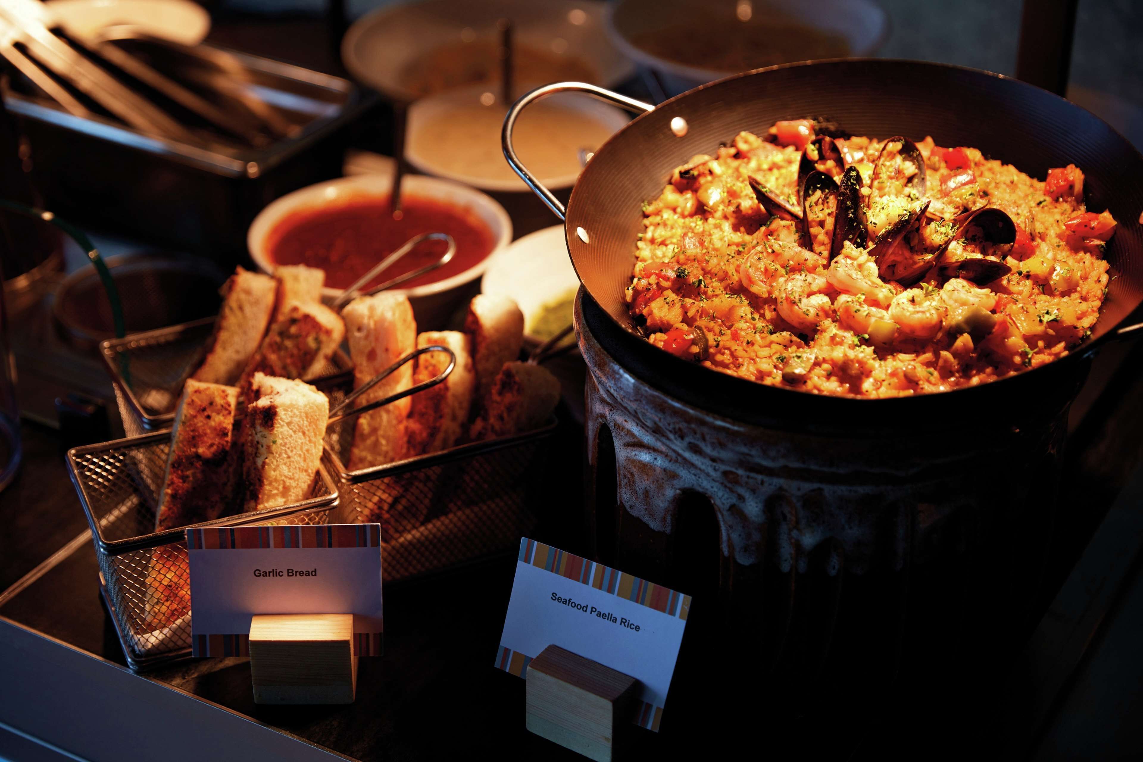 A seafood paella dish in a large pan, accompanied by garlic bread and a bowl of sauce.